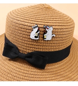 Detail of two cartoon cat enamel pins on a straw hat featuring a kitten drinking wine