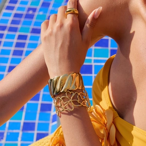 Close-up of model wearing gold hollow heart bangle and textured bracelets by a pool