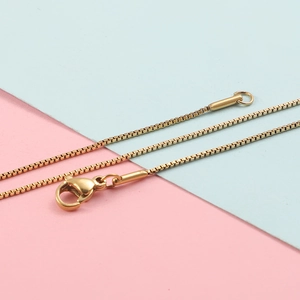 Detail van een gouden box chain ketting met karabijnsluiting op roze en blauwe achtergrond
