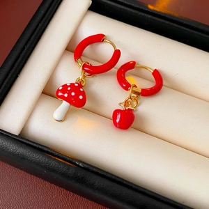 Macro shot of asymmetric mushroom and cherry red enamel flower earrings on a beige cushion