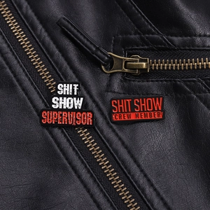 Detail view of funny Shit Show Crew enamel pins on black leather jacket textures