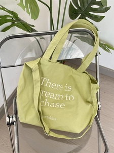 Light green canvas tote bag with lettering on a clear chair with plant background