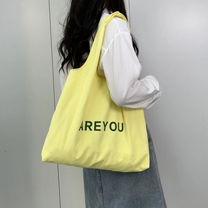 Detail of a yellow canvas tote bag with ARE YOU lettering worn by a model in a white shirt