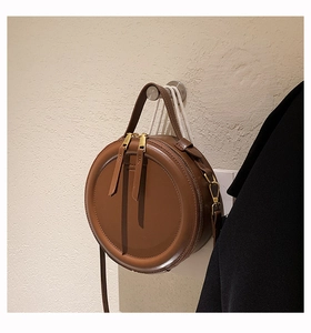 Detail einer runden braunen Ledertasche mit goldenen Beschlägen an einer schlichten Wand