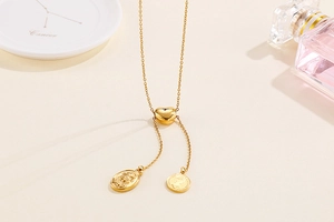 Detail of gold Y-shaped necklace with heart bead and engraved coin pendants on beige surface