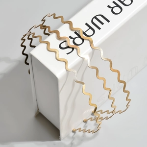 Three minimalist gold wave-shaped metal headbands displayed on a modern white pedestal
