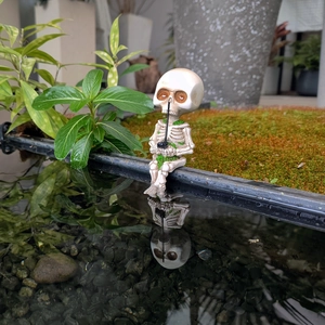 Detailed resin skeleton fishing figurine sitting by water with green moss and fishing rod