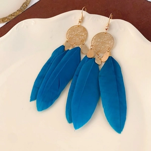 Detail of long peacock blue feather earrings with golden boho charms on a white porcelain tray