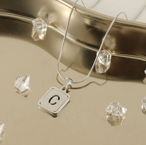 Detailed close-up of silver snake chain with matte square letter C pendant on gold tray