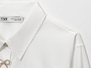 Dettaglio del colletto di camicia bianca in popeline con fiocco dorato e bottoni in perle