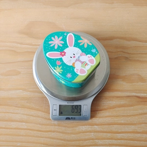Detailed top view of a bunny print egg-shaped tin box weighed at 69.3g on a kitchen scale
