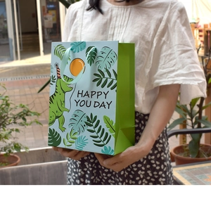 Handheld view of cartoon dinosaur paper gift bag with HAPPY YOU DAY text for kids