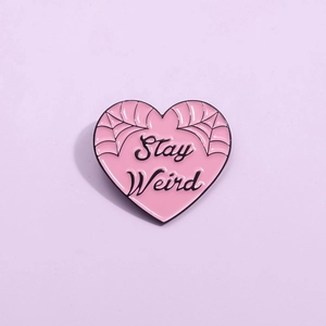 Pink heart-shaped enamel pin with spider web pattern and Stay Weird lettering