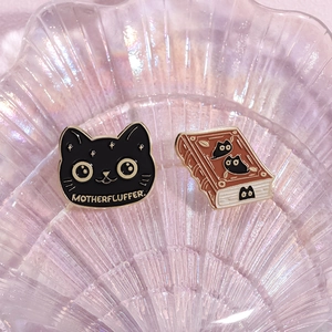 Close-up of enamel pins featuring a black cat and a magic book on a shell tray