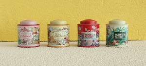 Row of four illustrated tin storage boxes in red, yellow, beige and pink finishes