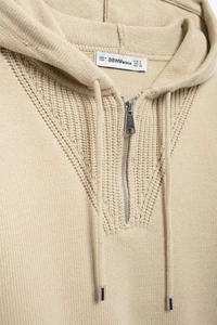 Detailed close-up of beige knitted hoodie neckline with half-zip and branded label