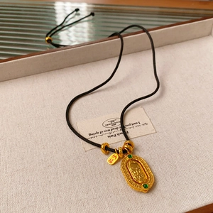 Macro detail of gold oval pendant with green gems and a Na Fu character tag on black cord