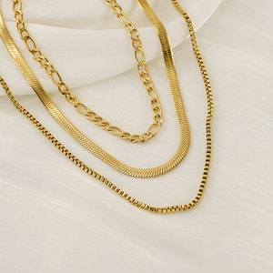 Layered gold snake, figaro, and box chains on cream fabric background for women