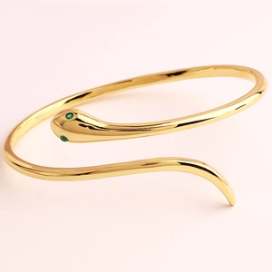 Close-up of golden snake bracelet featuring green gemstone eyes on soft pink background