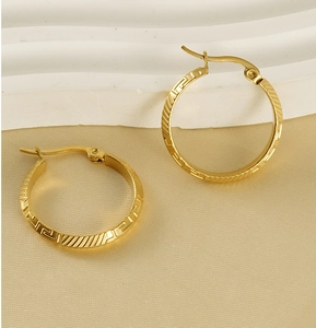 Detailed view of gold hoop earrings featuring an engraved Greek fret pattern on a beige surface