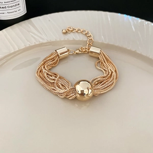 Detail of a layered gold chain bracelet with a large metal ball bead on white ceramic tray