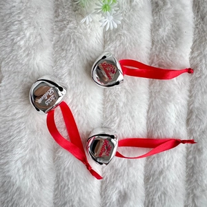 Three silver 2025 Believe Christmas bells with red ribbons on white plush background