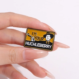 Cartoon enamel pin with I'M YOUR HUCKLEBERRY text and cowboy figure held by a hand