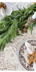 Close-up of lifelike PE green garland on vintage lace tablecloth for holiday dining decor