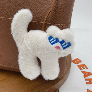 Close-up of a white plush cat keychain with blue heart eyes on a brown leather bag