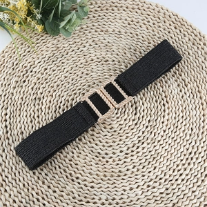 Detail of a black elastic woven belt featuring a textured gold metal buckle on a straw mat