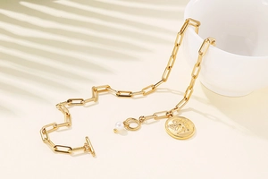 Golden paperclip chain necklace with angel coin pendant and pearl accent on a beige surface