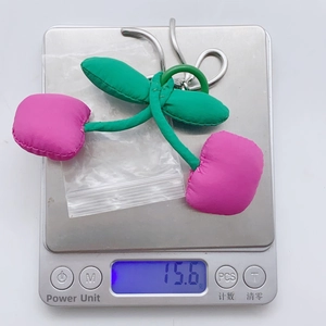 Detail showing pink fabric cherry keychain on digital scale weighing 15.6 grams