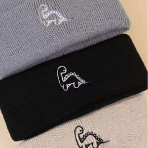 Detailed view of three knitted beanies in grey, black, and beige with dinosaur embroidery