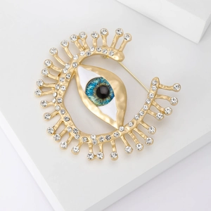 Broche œil grec dorée avec strass blancs et détail bleu sur un support géométrique blanc