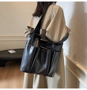 Large black PU leather tote bag with functional front pockets shown on a beige trench coat