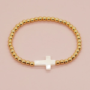 Handmade gold beaded bracelet with a white cross charm on a soft pink background