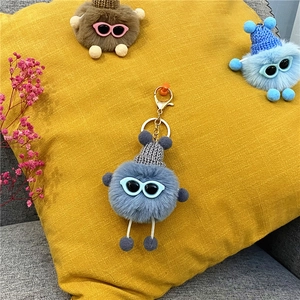 Blue fuzzy plush monster keychain with gray knit hat and glasses shown on a yellow cushion