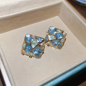 Macro detail of geometric square gold studs with blue and green inlay in a beige jewelry box