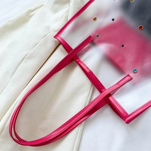 Transparent PVC tote bag with pink trim and colorful rivets on a beige fabric background
