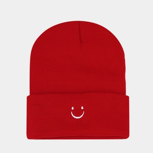Close-up of a red knitted beanie featuring a white embroidered smiley face and ribbed texture
