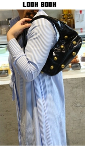Model wearing a black patent leather shoulder bag with gold rivets in a cafe setting