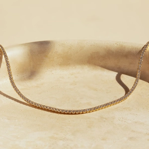 Close-up of golden tennis necklace with clear zircon stones on a beige stone tray