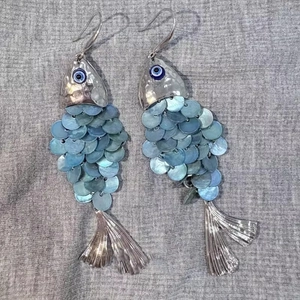 Detail of silver fish earring with blue shell scales and evil eye charm on fabric