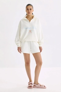 Détail du sweat à demi-zip crème avec écusson vintage et texture de broderie fine