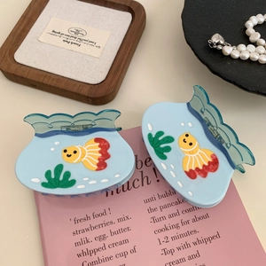 Blue fish tank hair claw clips with goldfish and seaweed patterns on pink book background