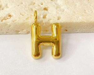 Macro shot of gold-plated 3D bubble letter H pendant on textured beige stone base