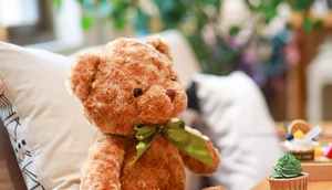 Close-up of a soft curly brown teddy bear plush with a green ribbon bow in a cozy setting