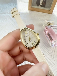 Detailed close-up of a teardrop-shaped gold watch with a white textured leather strap
