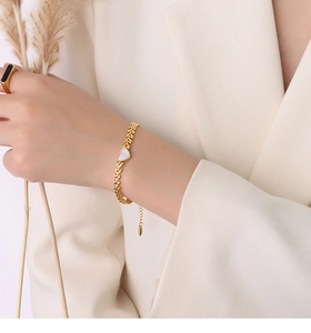 Detail of a 18K gold wheat chain bracelet with a white shell heart on a model wrist