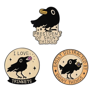 Three enamel crow pins with shiny trinkets and humorous quotes on white background